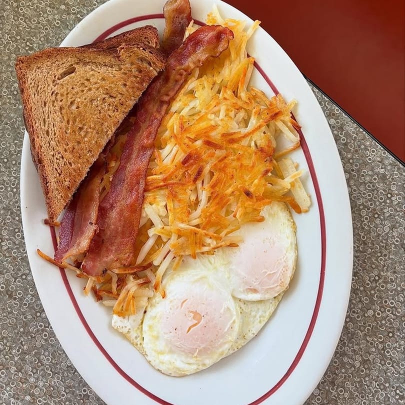 A diner breakfast plate with crispy bacon, hash browns, two fried eggs, and toast.