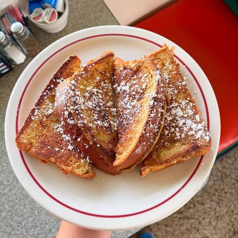 Four slices of golden-brown French toast, dusted with powdered sugar, on a white plate with a red rim.